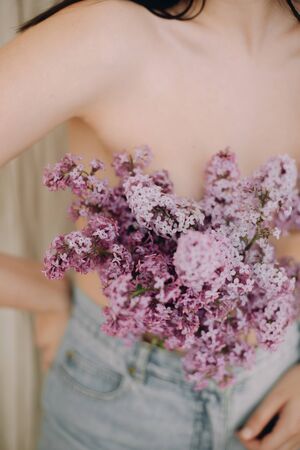 Woman posing with lilac branches in denim jeans on rustic background. Blooming lilac flowers covering naked upper body. Creative moody image. Sensuality and tenderness conceptの写真素材