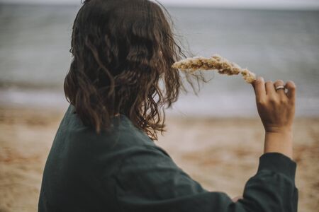 Carefree hipster girl with windy hair holding herb on beach. Stylish tanned boho woman in modern outfit relaxing on seashore. Summer vacation. Mindfulnessの写真素材