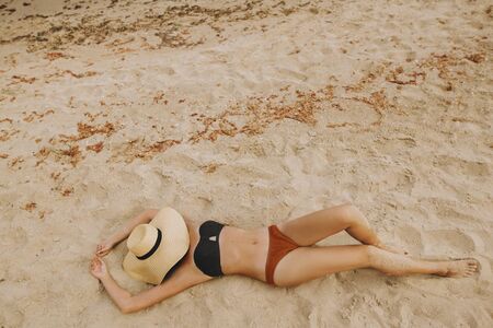 Fashionable young woman in straw hat relaxing on sandy beach near sea. Summer vacation and travel. Girl lying on beach with hand holding hat. Carefree. Top viewの写真素材