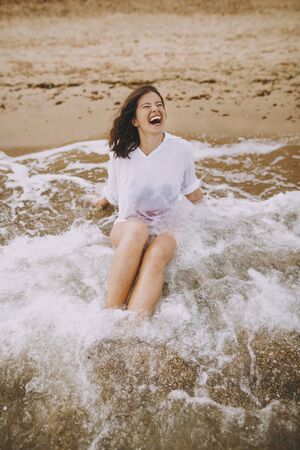Happy young woman in wet white shirt lying on beach in splashing waves.  Emotional tanned girl...
