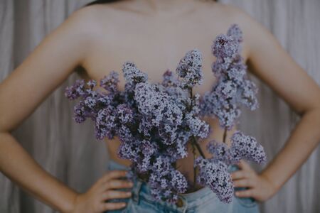 Woman posing with lilac branches in denim jeans on rustic background. Blooming lilac flowers covering naked upper body. Creative moody image. Sensuality and tenderness conceptの写真素材