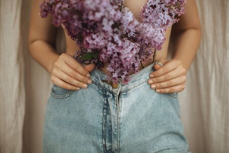 Woman posing with lilac branches in denim jeans on rustic background. Blooming lilac flowers...