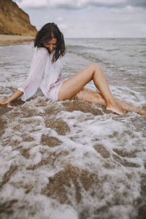 Happy  woman in white shirt sitting on beach in splashing waves. Stylish tanned girl relaxing on seashore and enjoying waves. Summer vacation. Mindfulness and carefree momentの写真素材