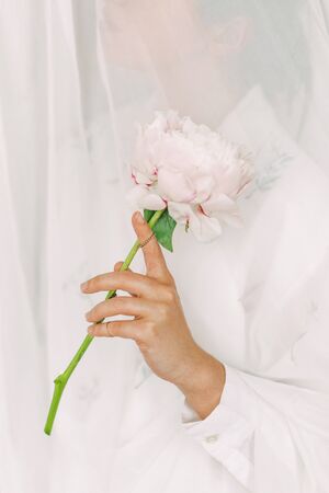 Aesthetic sensual image of beautiful woman holding pink peony flower through tulle. Stylish girl behind white textile gently holding peony flower in hand with jewelry. Soft imageの写真素材