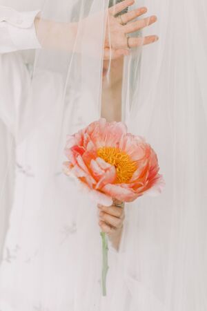 Aesthetic sensual image of beautiful woman behind veil holding pink peony. Stylish girl under tulle gently holding peony flower in hand with jewelry. Soft imageの写真素材