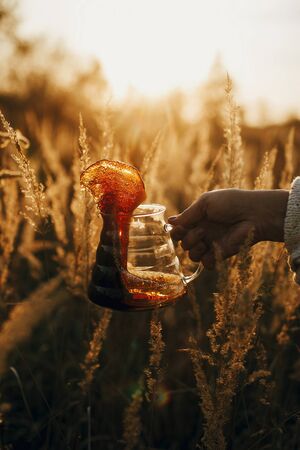 Coffee splash and drops in sun. Hand spilling out fresh hot coffee from glass flask in sunny warm light in rural herbs. Alternative coffee brewing in travel. Vertical imageの写真素材