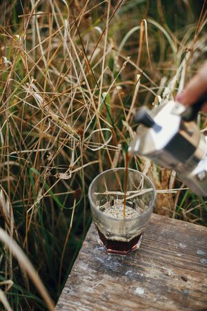 Alternative coffee brewing in travel. Pouring fresh hot coffee from geyser coffee maker into glass cup in sunny warm light in rural countryside herbs. Atmospheric moment. Vertical imageの写真素材