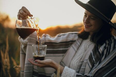 Hipster woman in hat pouring fresh hot coffee in glass cup of sunny warm light in rural herbs.の写真素材