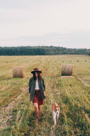 Stylish girl walking with her dog at hay bale in summer field in sunset. Young woman in hat traveling with pet in haystack field. Countryside slow lifeの写真素材