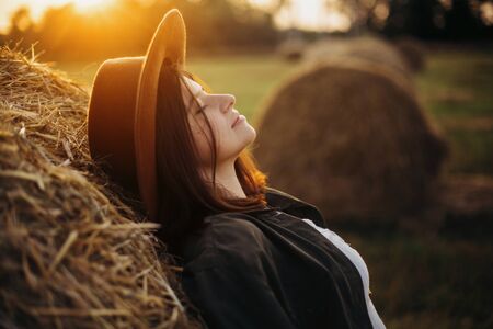 Stylish girl relaxing on hay bale in summer field in sunset. Portrait of young sensual woman in resting at haystack, atmospheric tranquil moment. Countryside slow lifeの写真素材