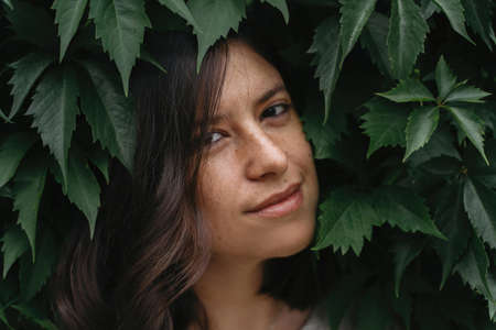 Sensual portrait of young brunette woman among greenery in park or garden. Calm moment. Stylish millennial girl standing in green leaves in city street.の写真素材