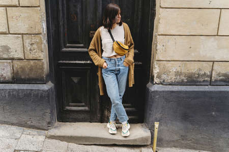 Stylish hipster girl standing at building in european city street. Young woman in casual fashionable outfit posing at old wooden door. Alone leisure activity in cityの写真素材