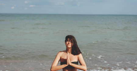 Young beautiful woman practicing yoga on the beach, sitting on sand and meditaning. Mental health and self care concept. Happy girl relaxing on seashore on summer vacationの写真素材