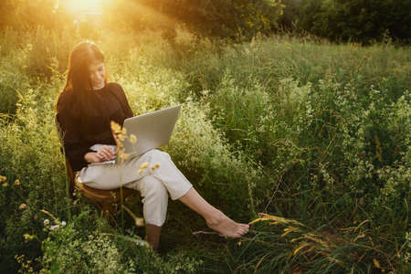 New office concept. Fashionable elegant girl working on laptop and sitting on rustic chair in warm sunset light in summer field. Remote work with social distancing and safety protocolsの写真素材
