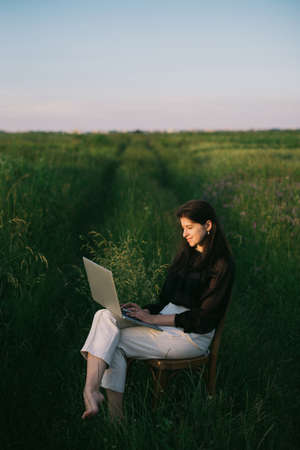 Young business woman working online. Freelance and remote work outdoors. Creative image. Fashionable elegant girl working on laptop and sitting on rustic chair  in summer field at sunset.の写真素材