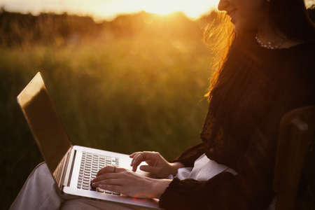 Freelance and remote work outdoors. Fashionable elegant girl with hands on keyboard sitting in sunny summer field at sunset.Young business woman working online on laptop. Creative imageの写真素材