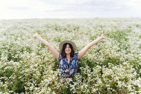 Happy young woman smiling in white wildflowers in summer meadow. Beautiful joyful girl in blue vintage dress and hat having fun in field, moment of happinessの写真素材