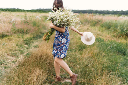 Beautiful girl having fun and laughing in windy field with big daisies bouquet. Authentic summer in countryside. Young woman in blue vintage dress and hat dancing with white wildflowers in meadow.の写真素材