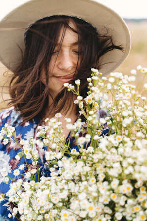 Sensual portrait of beautiful girl with big daisies bouquet in windy field. Summer in countryside. Beautiful young woman in blue vintage dress and hat holding white wildflowers in meadow.の写真素材