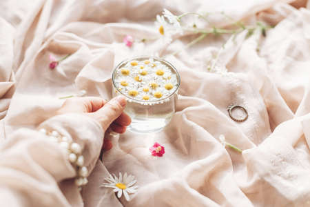 Hand with jewelry holding glass cup with daisy flowers in water on background of soft beige fabric with wildflowers. Tender floral aesthetic. Creative summer image. Bohemian moodの写真素材