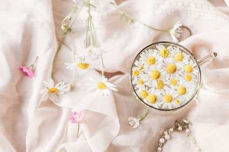 Tender floral aesthetic. Daisy flowers in water in glass cup on background of soft beige fabric with wildflowers and jewelry. Creative summer image, flat lay. Bohemian moodの写真素材