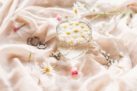 Tender floral aesthetic. Daisy flowers in water in glass cup on background of soft beige fabric with wildflowers and jewelry. Creative summer image with space for text. Bohemian moodの写真素材