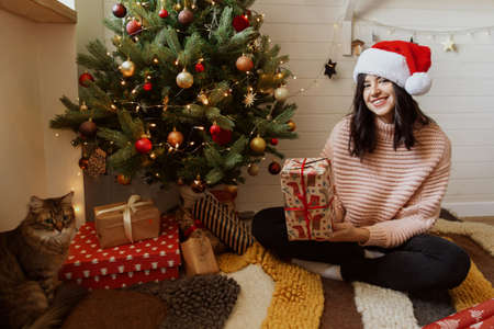 Stylish young woman holding red christmas gift box under christmas tree with lights in modern room. Happy girl in santa hat and sweater preparing christmas presents. Happy holidaysの写真素材