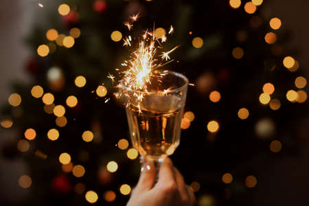 Happy New Year. Burning firework in champagne glass in hand on background of golden bokeh lights in festive room. Hand holding drink with sparkler at christmas tree. Creative aesthetic momentの写真素材