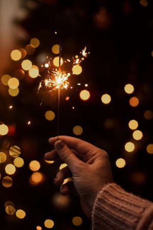 Burning sparkler in hand on background of golden bokeh lights in festive dark room. Happy New Year. Hand holding firework at christmas tree with golden illumination.の写真素材