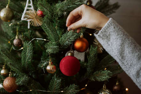 Woman decorating christmas tree with red bauble closeup. Preparation for christmas time. Modern simple ornament in hands on background of festive tree in lights. Happy holidaysの写真素材