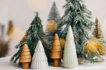Merry Christmas. Christmas trees and golden lights bokeh on white background. Winter magic forest, festive modern decor. Miniature ceramic, wooden, snowy and handmade pine trees.の写真素材
