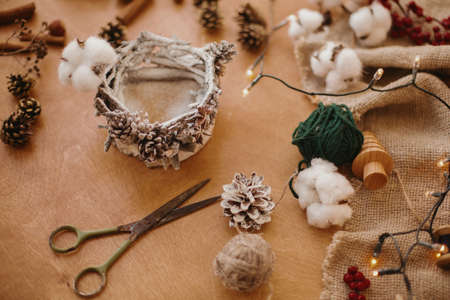 Making rustic christmas candle decor. Christmas candle  holder made of branches, cotton, anise and pine cones on wooden table with festive lights. Holiday workshop.の写真素材