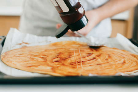 Round dough with ketchup for pizza. Person smearing tomato sauce on raw dough on modern white...