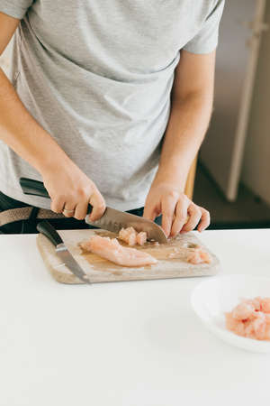 Person cutting chicken fillet with knife on wooden board on modern white kitchen. Process of making...