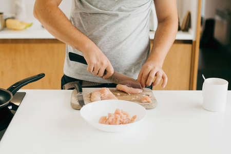 Person cutting chicken fillet with knife on wooden board on modern white kitchen. Process of making home pizza, hands close up. Home cooking conceptの写真素材