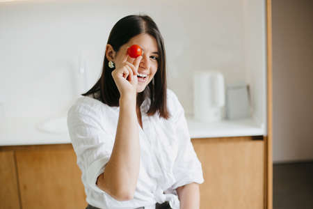 Young happy woman holding cherry tomato and smiling on background of modern white kitchen. Healthy food concept. Home cooking, salad ingredientsの写真素材
