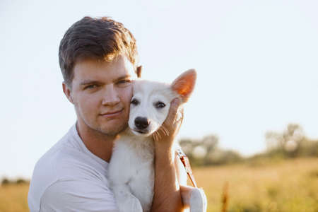 Young handsome man hugging cute white puppy in warm sunset light in summer meadow. Person caressing adorable fluffy puppy.Traveling with dog or adoption concept, loyal friendの写真素材