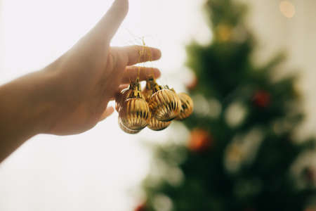 Decorating christmas tree. Hand holding modern gold baubles on background of green tree. Hanging ornaments on christmas tree, festive decoration of roomの写真素材