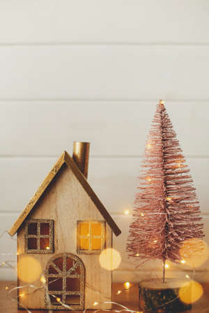 Stylish christmas house and glitter christmas tree in golden lights. Modern festive decor in scandinavian room. Miniature village with wooden house and tree toys. Merry Christmasの写真素材