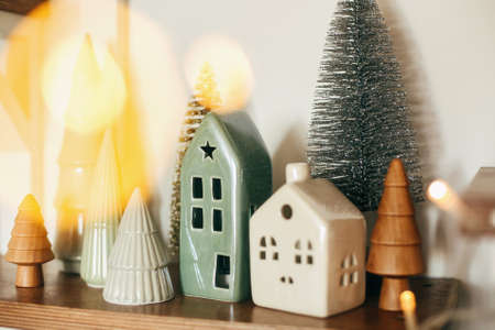 Modern little christmas ceramic house and glitter trees on wooden shelf, stylish festive decor....