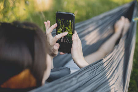 Young woman chilling in hammock and taking photo of her legs. Millennial girl holding phone and...