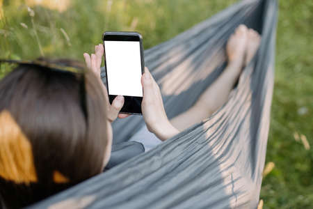 Young woman chilling in hammock and holding phone with empty screen. Girl with phone chatting with friends or making order online, while relaxing in hammock. Phone mock upの写真素材