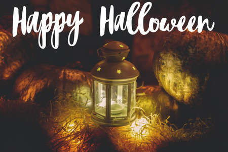 Happy Halloween greeting card. Hand written Happy Halloween text on background of glowing lantern on hay bale and golden pumpkins in darkの写真素材