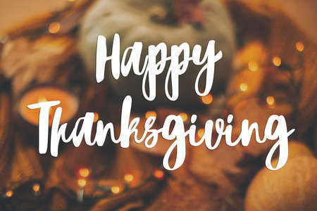 Happy Thanksgiving greeting card. Hand written Happy Thanksgiving text on blurred background of pumpkin, autumn leaves, warm lights on yellow knitted sweater.の写真素材