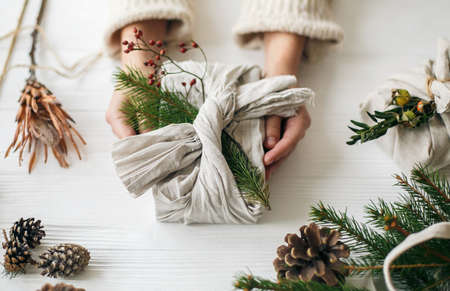 Zero waste Christmas holidays. Hands holding stylish gift wrapped in linen fabric with green fir branch and red berries on rustic white table with pine cones. Plastic free holidaysの写真素材