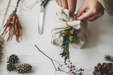 Female preparing zero waste christmas present. Hands decorating stylish christmas gift in linen fabric with green branch on white rustic table with natural herbs and pine cones.の写真素材