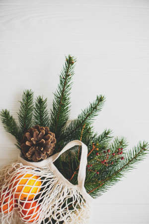 Net cotton bag with green spruce branches, oranges and pine cones on white rustic background. Reusable shopping bag with winter decorations, plastic free holidays. Top viewの写真素材