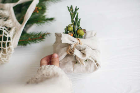 Hand holding stylish christmas gift wrapped in linen fabric with green  branch on rustic white wooden background. Zero waste christmas holidays. Sustainable lifestyleの写真素材