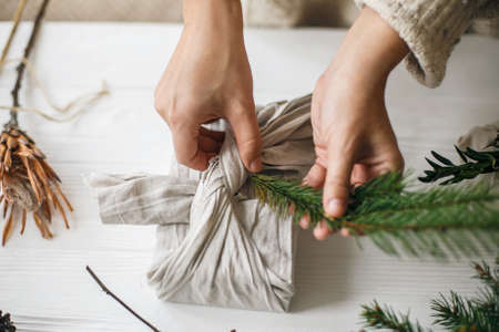 Hands decorating stylish christmas gift in linen fabric with green fir branch on white rustic table with pine cones and berries. Florist preparing zero waste Christmas gift. Plastic free holidaysの写真素材