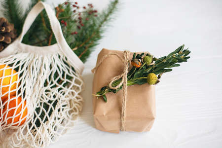 Stylish christmas gift box wrapped in craft paper with natural green branch on background of reusable cotton bag with oranges and fir branches on white table. Zero waste christmas holidaysの写真素材